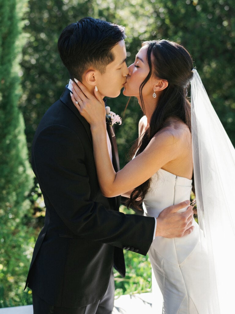 Romantic bride and groom sharing a passionate kiss outdoors, highlighting an editorial bridal look and classic black tuxedo during their destination wedding travel.
