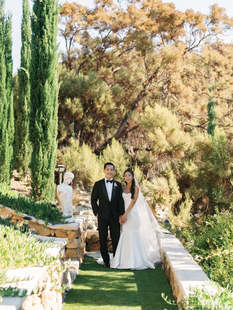 Chic bride and groom posing on a lush garden path flanked by tall cypress trees during their luxury destination wedding travel at a California estate.