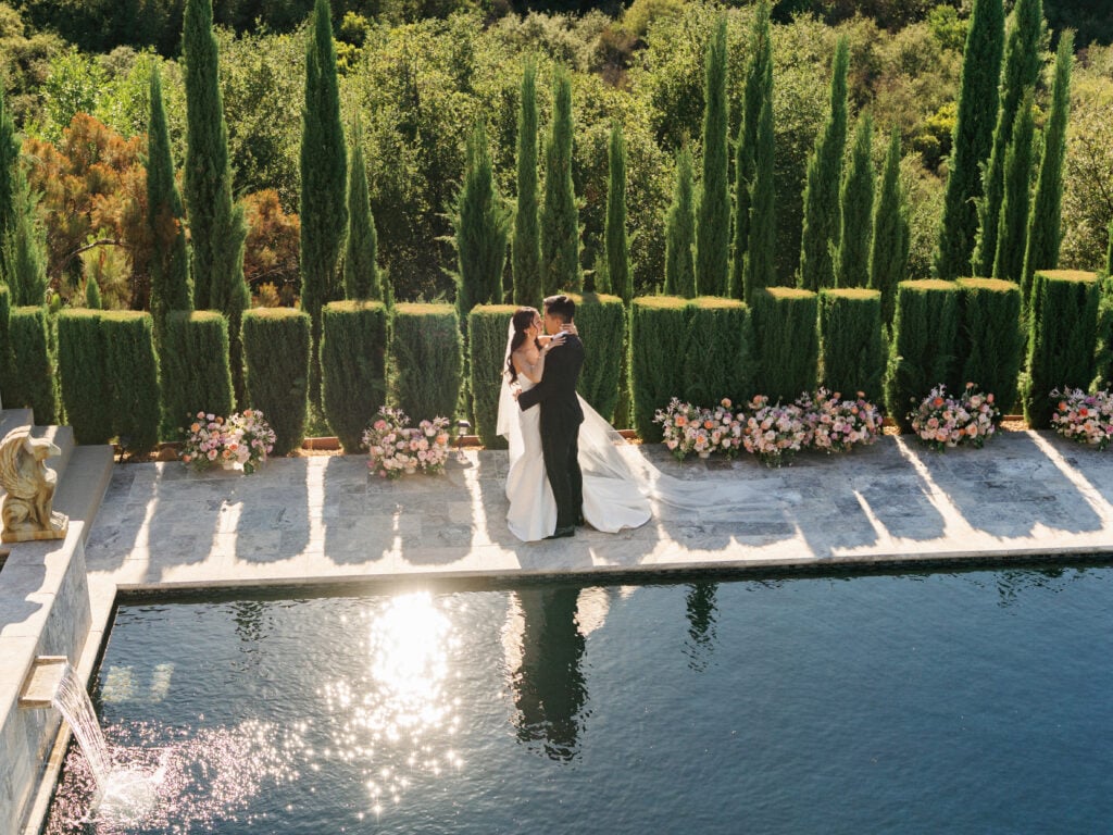 Bride and groom embracing by a luxury infinity pool with sun reflections and tall cypress trees, capturing high-end destination wedding travel design inspiration.