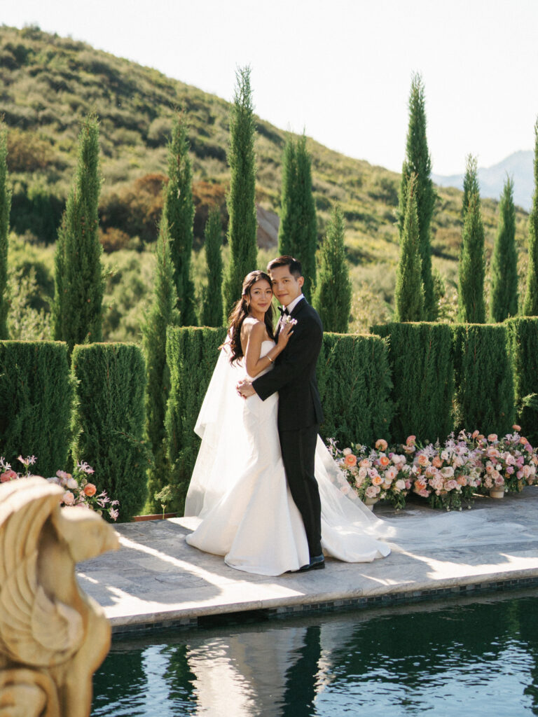 Luxury wedding photo ideas featuring an elegant couple posing by the pool at Sunstone Winery villa surrounded by cypress trees.