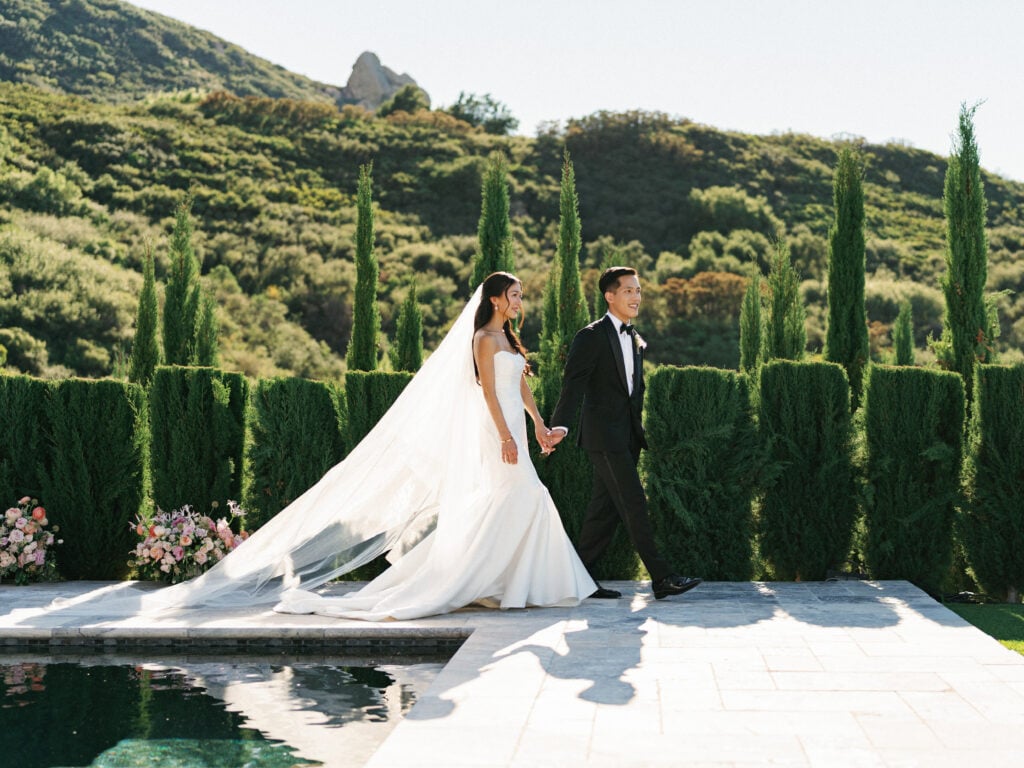 Elegant bride in a sleek satin mermaid gown with a flowing cathedral veil walks hand-in-hand with her groom by a luxury poolside terrace during their destination wedding travel in Malibu.