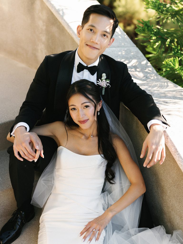 Bride in strapless gown and groom in tuxedo seated together on outdoor stone steps.