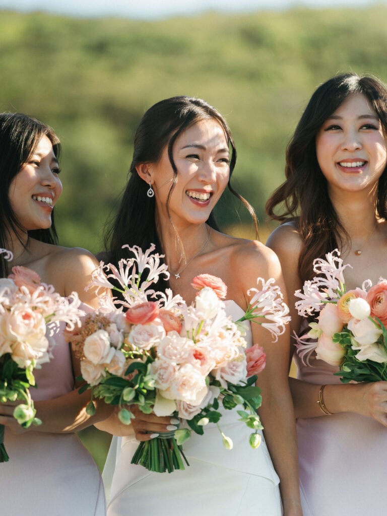 Laughing bride and bridesmaids holding vibrant peach and pink bouquets during an outdoor luxury destination wedding travel celebration in Malibu.