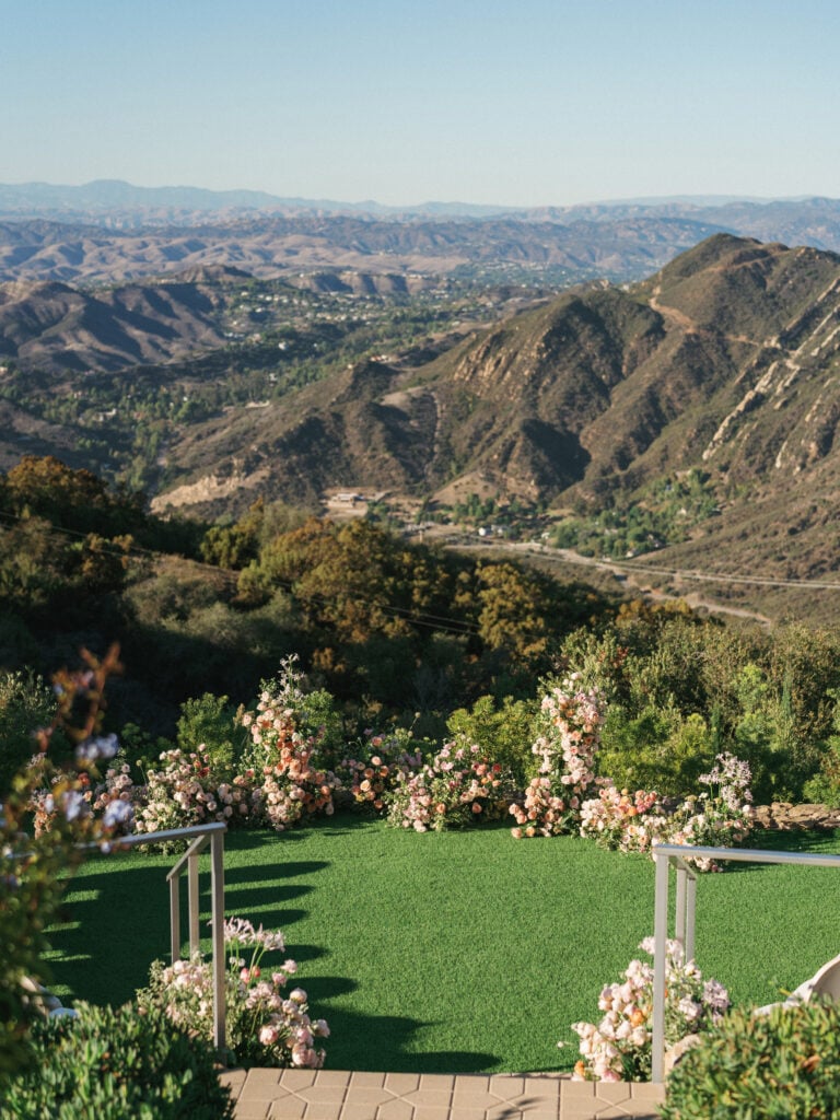 Luxury outdoor wedding ceremony setup on a green lawn with blush floral installations overlooking the breathtaking Malibu canyon mountains during destination wedding travel.