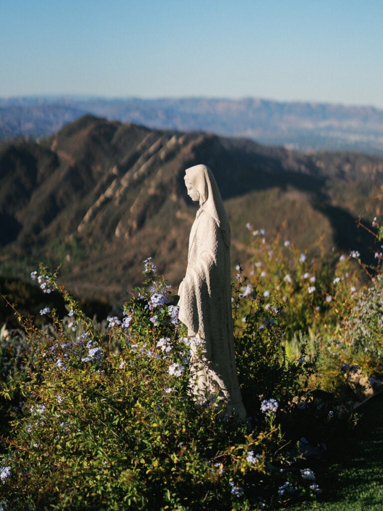 Classical stone Virgin Mary statue surrounded by wild shrubs overlooking a panoramic mountain view during a luxury destination wedding travel event in Malibu.