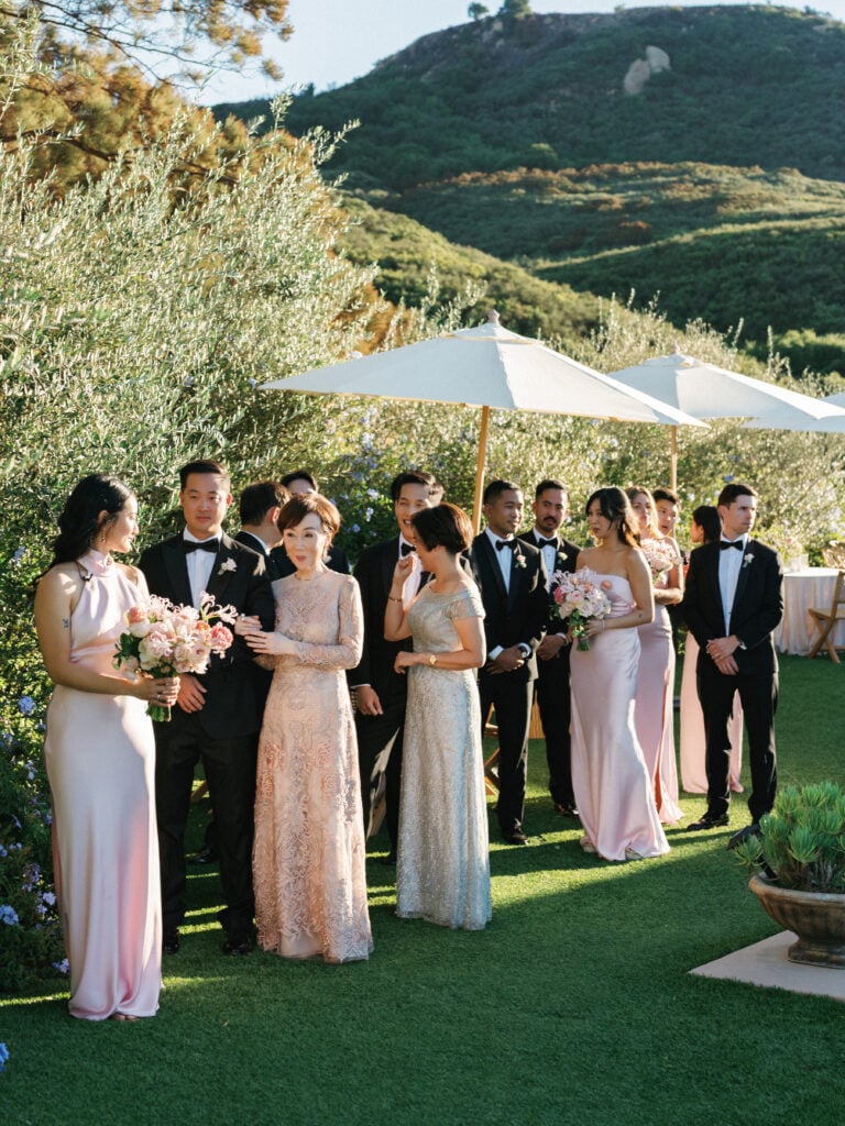 Wedding guests and bridal party in elegant blush pink and grey dresses mingling under umbrellas during a luxury destination wedding travel event in Malibu.