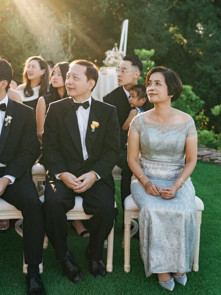 Parents of the bride and groom watching the ceremony in sophisticated formal attire on a sunny outdoor terrace during a high-end destination wedding ceremony.