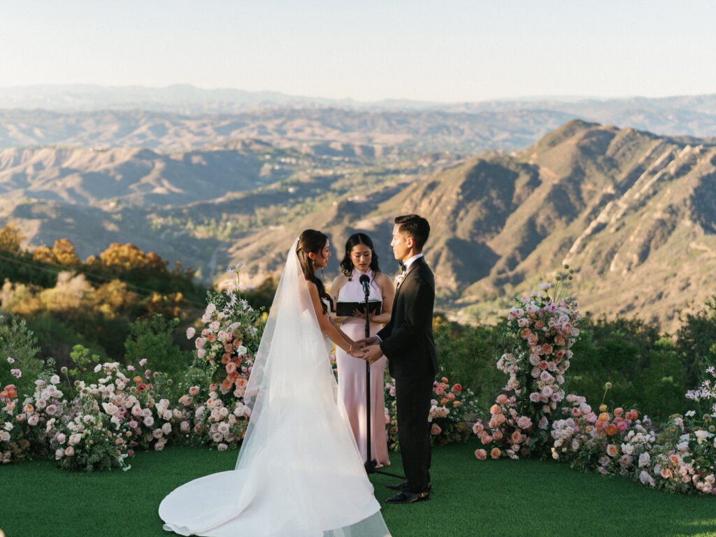 Bride and groom holding hands during a luxury outdoor wedding ceremony framed by lush pink floral arches overlooking the Malibu mountains, ideal inspiration for destination wedding travel.