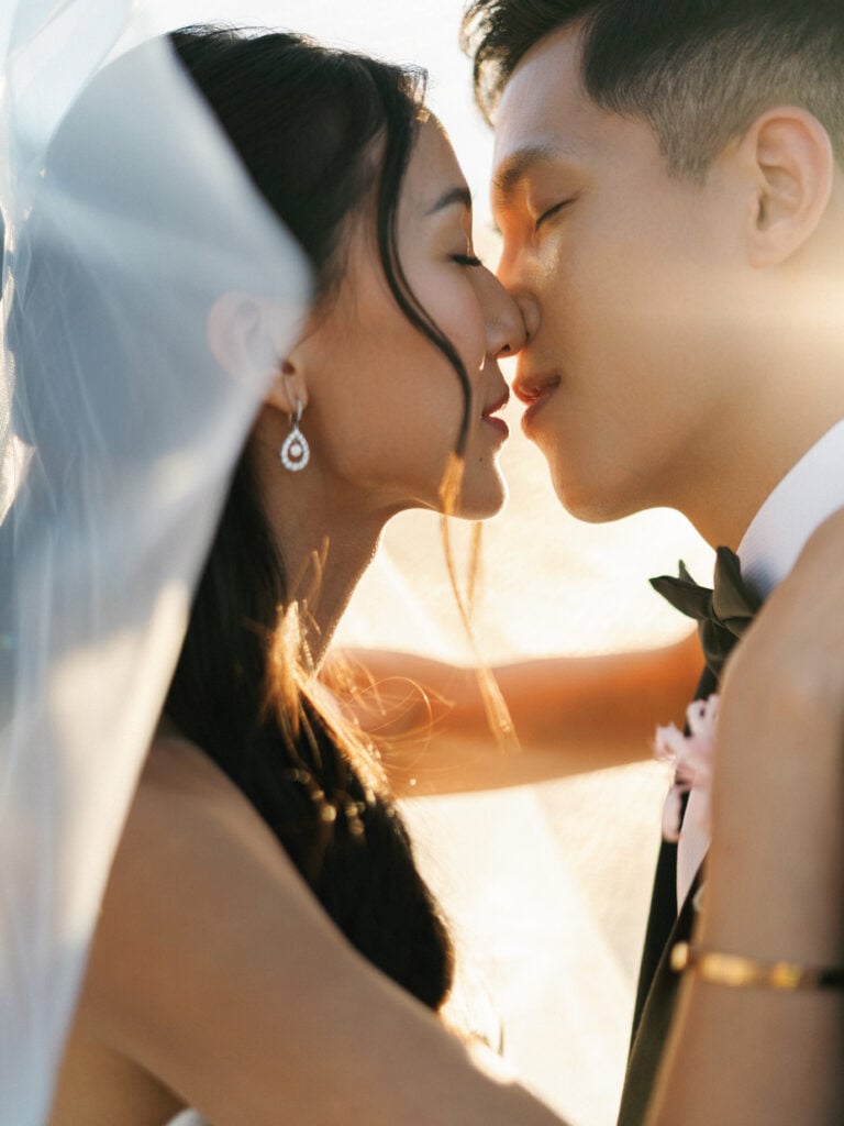 Close-up of a romantic bride and groom kissing in warm golden hour sunlight under a flowing veil, showcasing cinematic destination wedding travel photography.