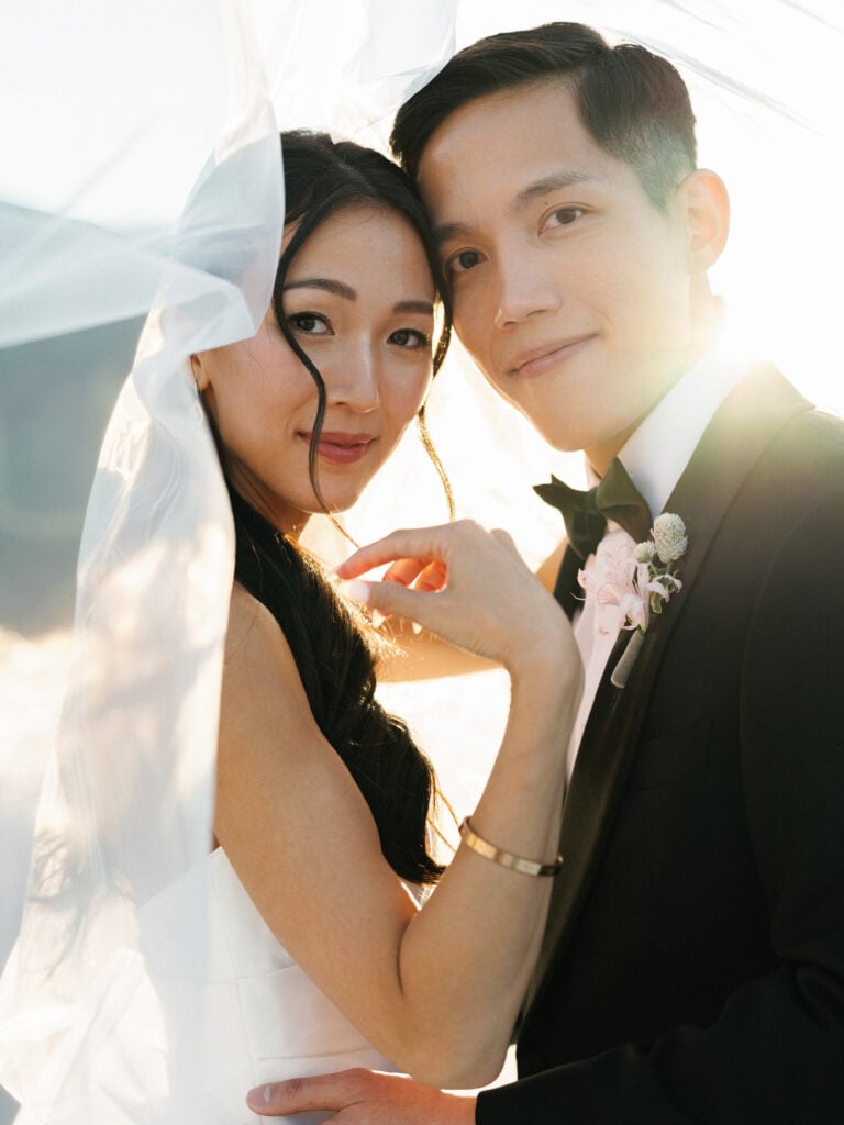 Elegant bride and groom sharing a close, tender look enveloped in a flowing cathedral veil during a glowing golden hour shoot, emphasizing natural emotion for editorial wedding photography.