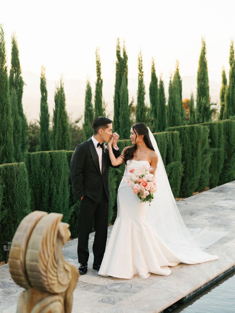 Groom kissing the bride's hand on a stone poolside terrace surrounded by manicured cypress trees during their luxury destination wedding travel in Malibu.