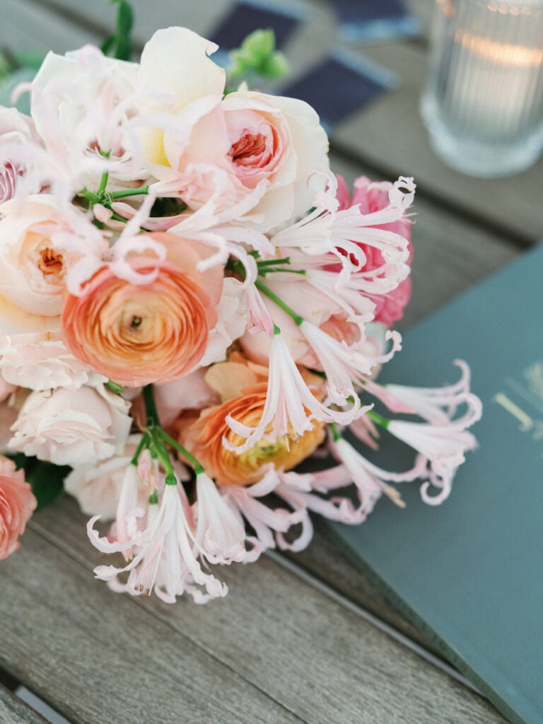 Close-up of an elegant bridal bouquet with peach ranunculus and pink nerine lilies on a wooden table, perfect details for destination wedding travel inspiration.