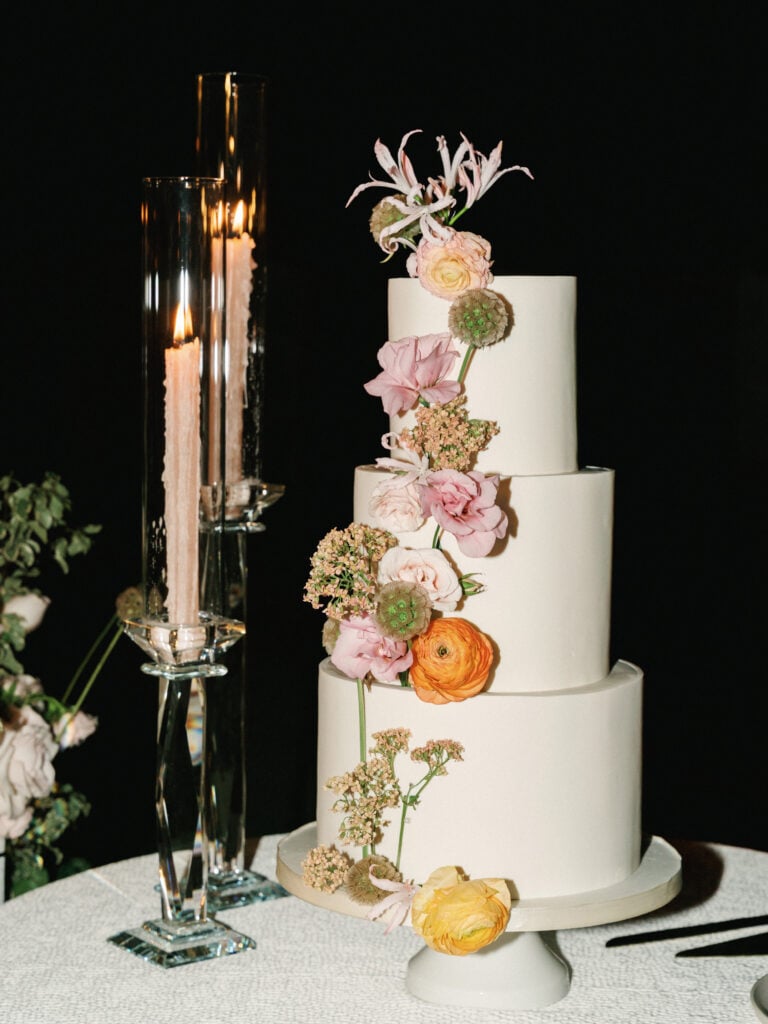 Three-tier white wedding cake adorned with fresh pastel flowers and tall glass taper candles on a reception table, perfect inspiration for luxury destination wedding travel.