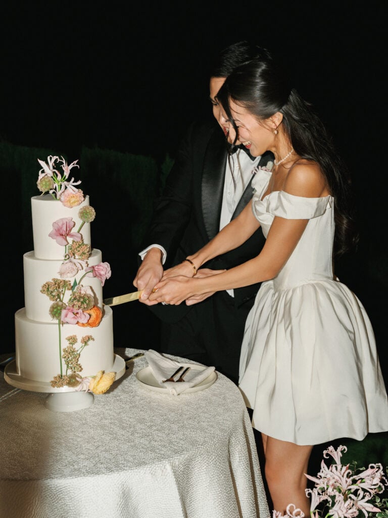 Smiling bride in a short satin reception dress and groom cutting their tiered floral wedding cake at an outdoor night reception during their destination wedding travel.