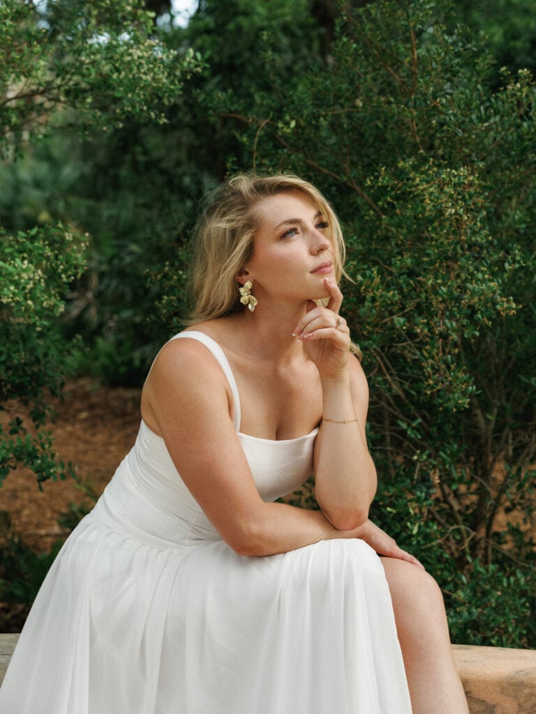 Thoughtful bride in a minimalist square-neck wedding dress with gold floral statement earrings posing in a lush garden during an editorial wedding photography session.