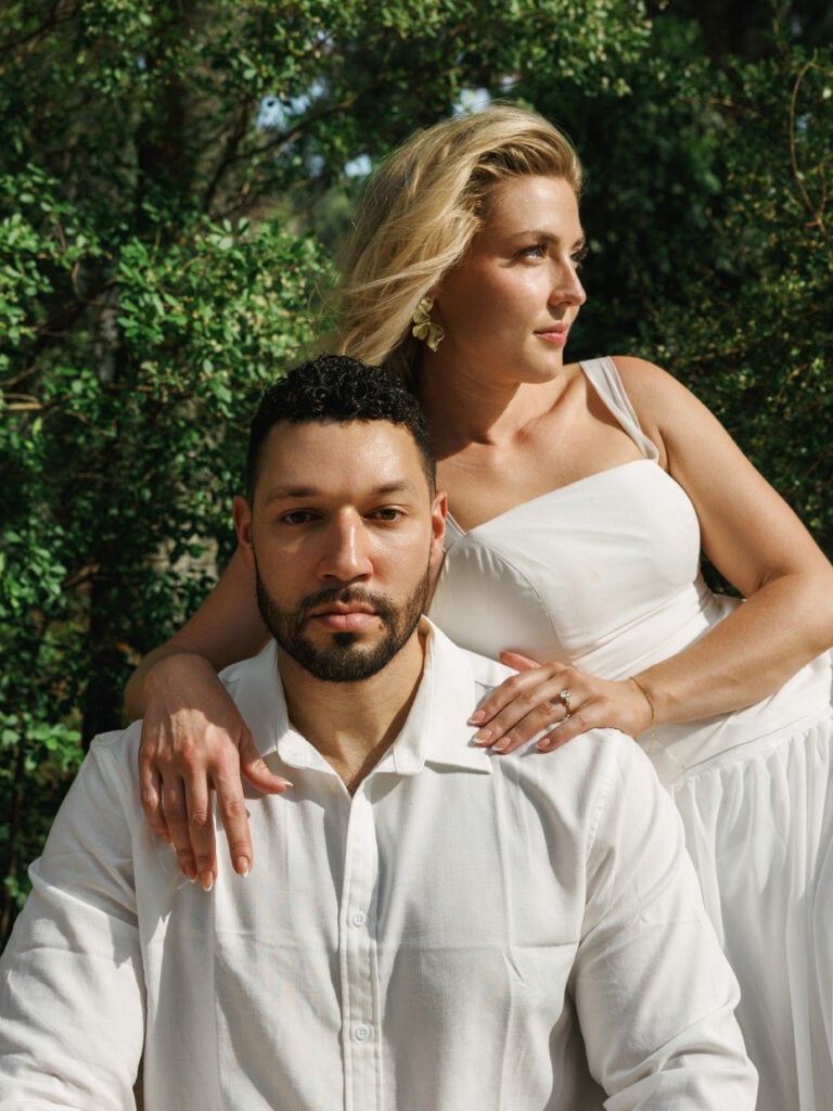 Elegant bride leaning on her groom's shoulders as they pose naturally in dappled sunlight under green trees, capturing modern editorial wedding photography style.