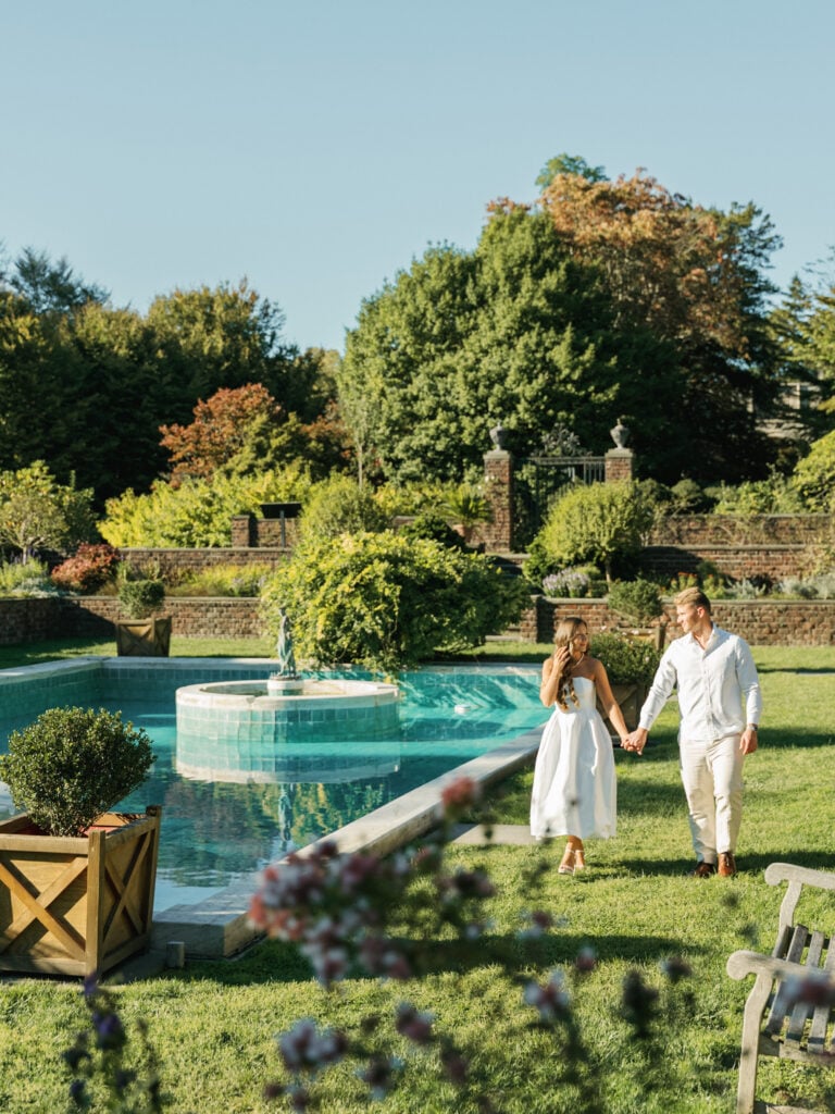 Chic bride in a strapless midi wedding dress walking hand-in-hand with her groom past an elegant turquoise pool with a stone fountain at a luxury estate, capturing modern editorial wedding photography.