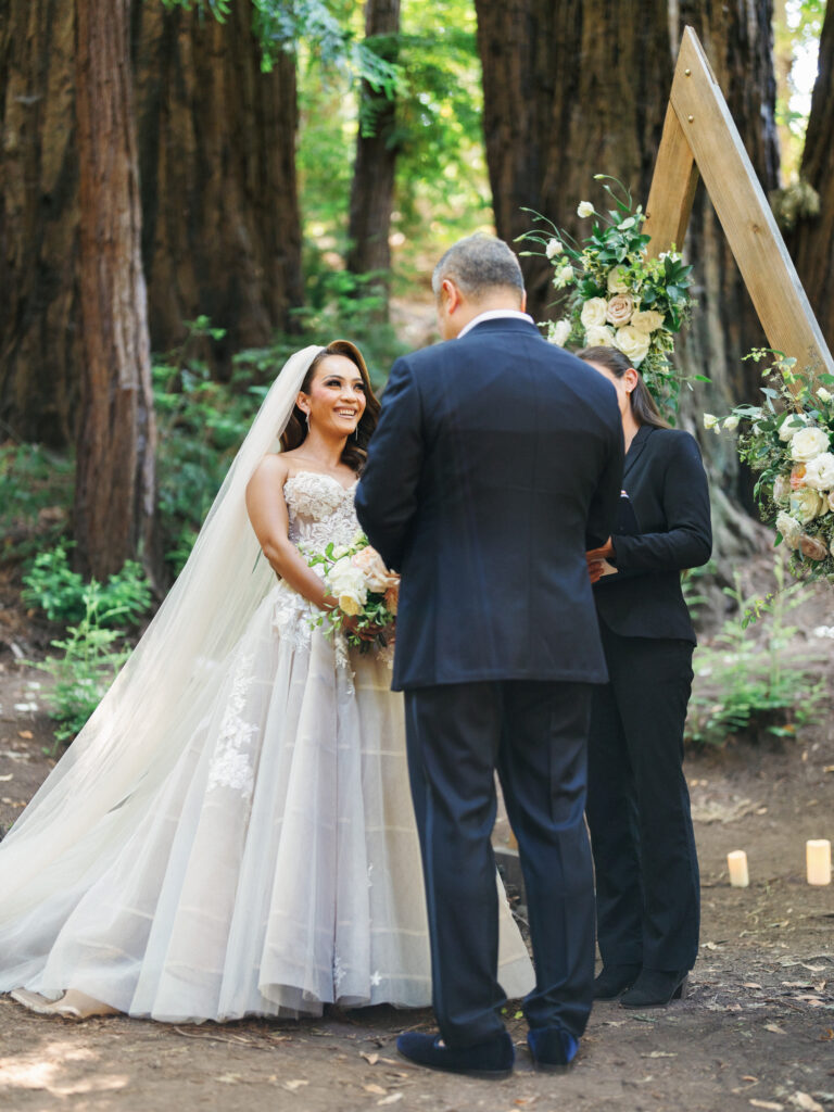 Bride and groom exchanging vows in front of a wooden triangle arch with flowers in a redwood forest