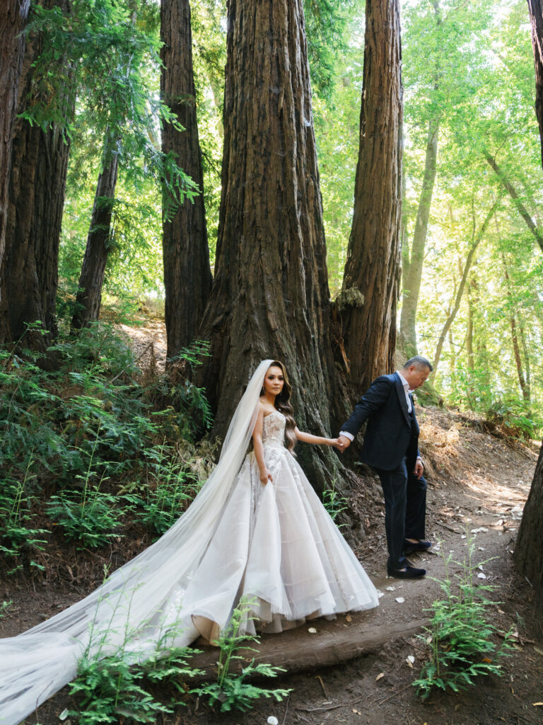 Bride in a full tulle gown holding hands with her groom while walking along a path between giant redwood trees captured by a Big Sur wedding photographer