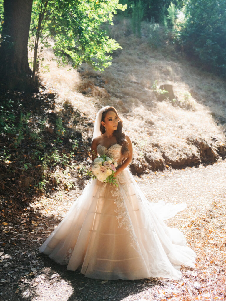 Bride in a tulle applique gown standing on a sunlit dirt path surrounded by trees in Big Sur California