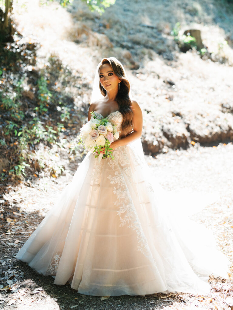 Bride holding a pastel flower bouquet while standing on a sunlit forest trail captured by a Big Sur wedding photographer