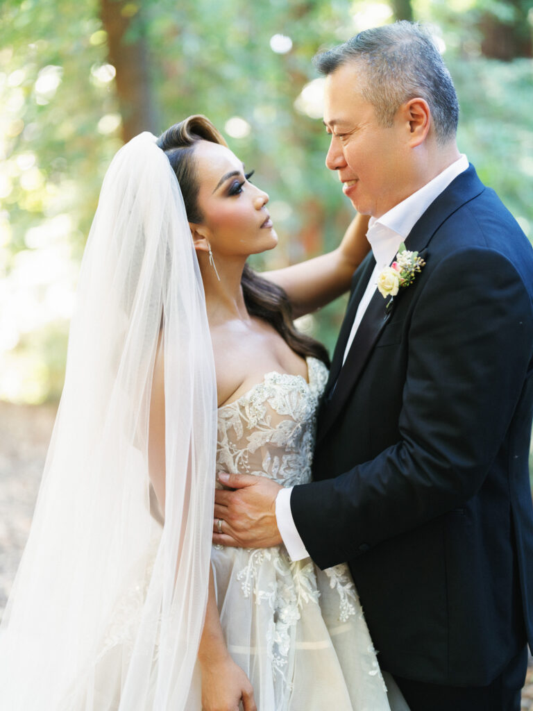 Bride with a long veil looking into her grooms eyes while standing in a sunlit forest in Big Sur California
