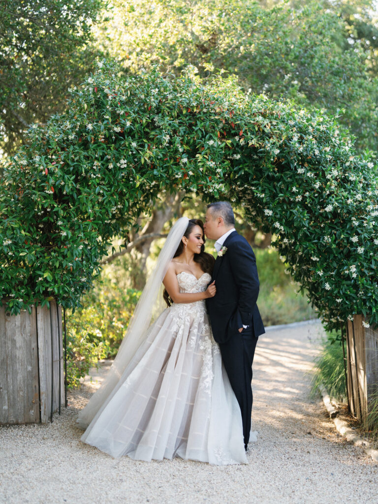Groom kissing his bride on the forehead while standing under a lush green garden arch captured by a Big Sur wedding photographer