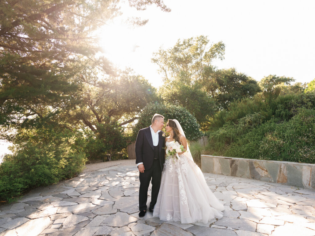 Bride and groom holding hands on a stone terrace with a dramatic sunset flare captured by a Big Sur wedding photographer