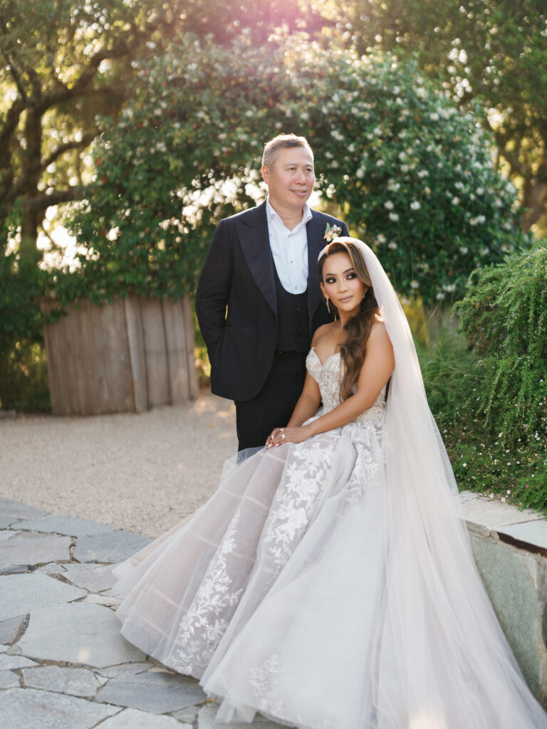 Bride in a flowing tulle gown with a long veil sitting on a stone ledge next to her groom in a tuxedo