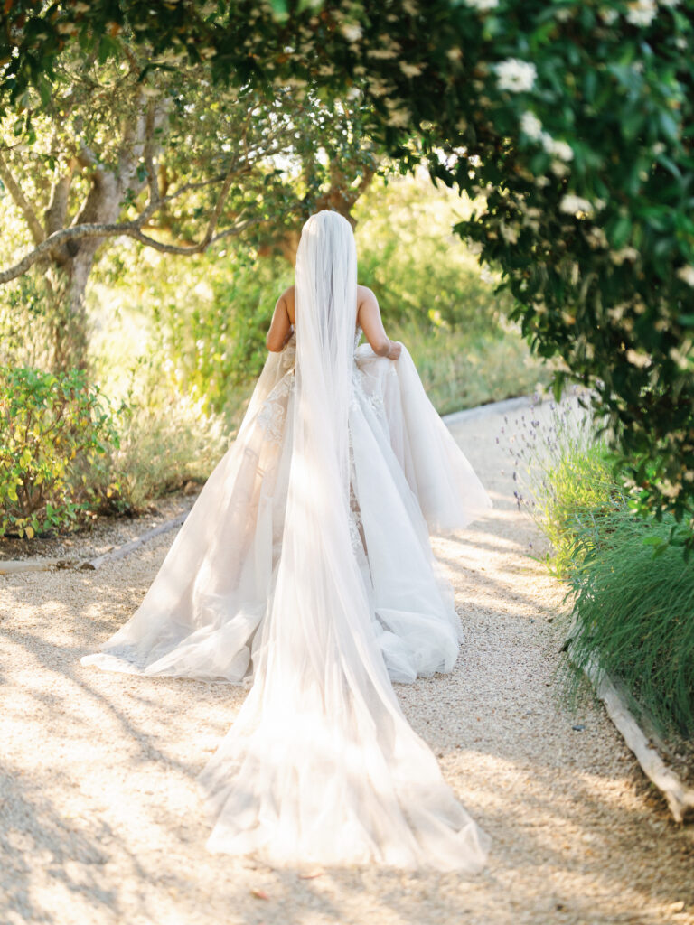 Bride walking away on a gravel path showcasing a long cathedral veil and wedding dress train under sunlit trees
