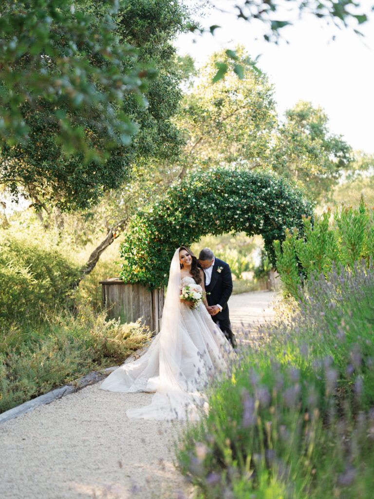 Newlyweds strolling under a green floral arch past lavender bushes captured by a Big Sur wedding photographer