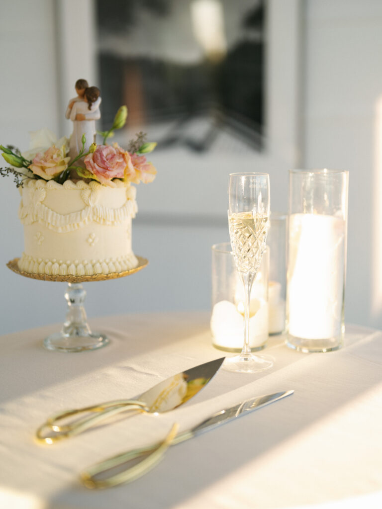 Single tier wedding cake with a topper next to a champagne flute and candles on a white tablecloth during sunset