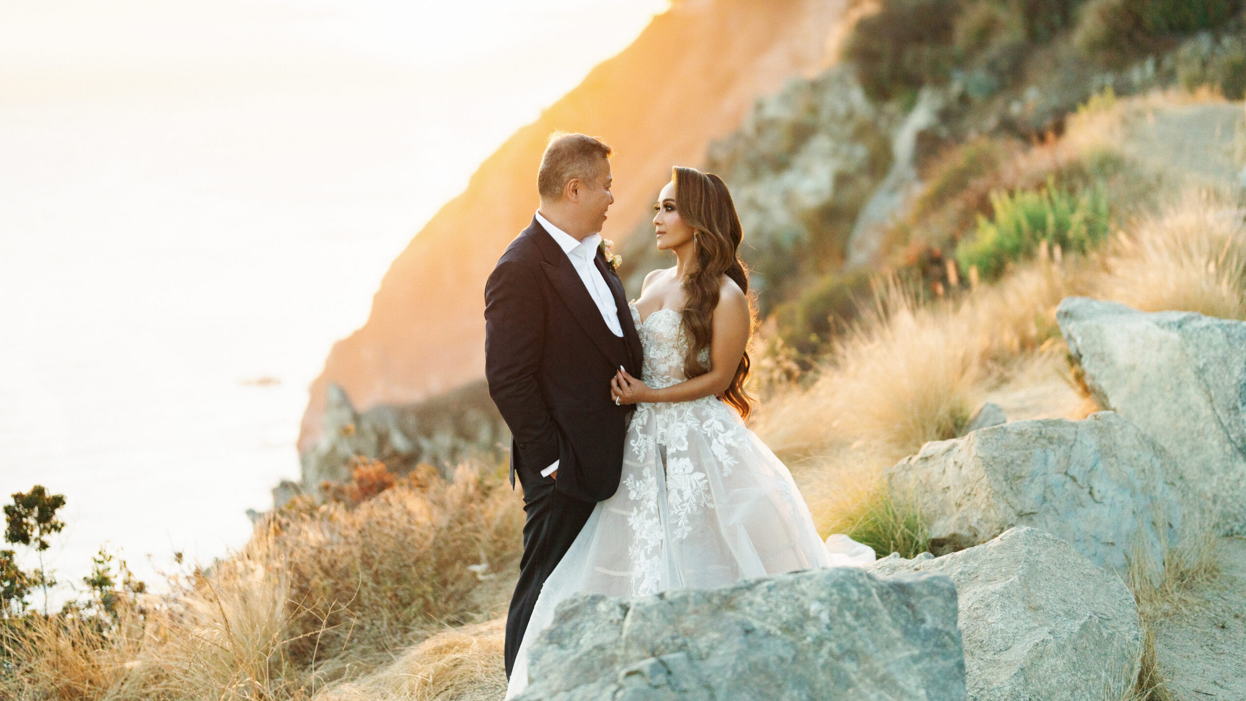 Newlyweds posing on a steep coastal hillside with golden dry pampas grass and rocky terrain in Big Sur California