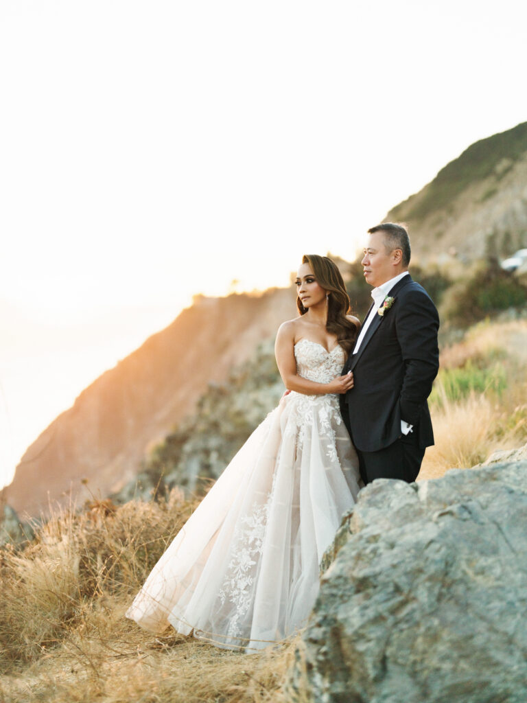 Bride and groom posing together on a rocky coastal hillside with a glowing sunset background captured by a Big Sur wedding photographer