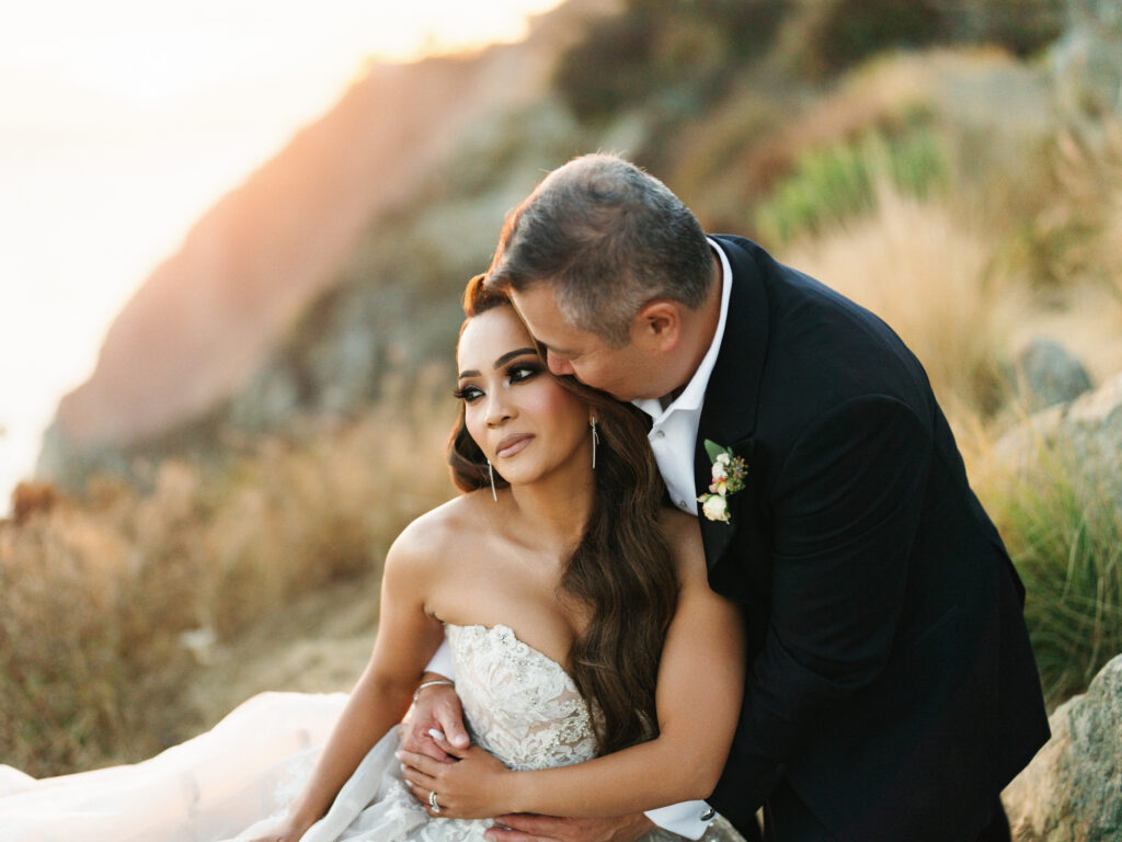 Groom embracing and kissing his smiling bride on the cheek during a sunset cliffside photoshoot in Big Sur California