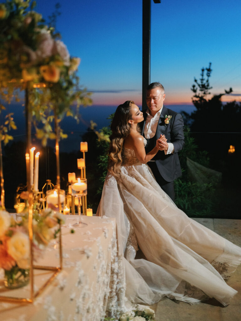 Bride and groom dancing on a terrace next to a decorated dinner table with a sunset sky background captured by a Big Sur wedding photographer