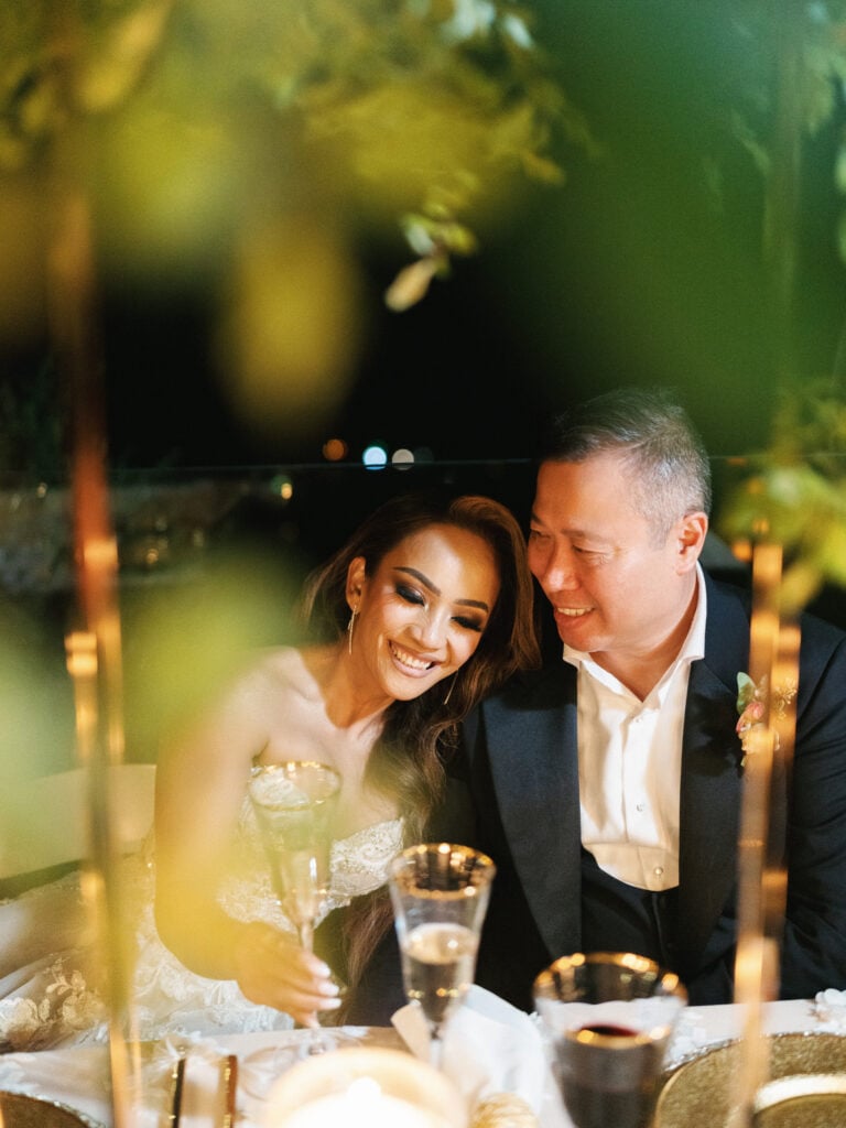 Bride and groom laughing at a candlelit head table framed by gold floral stands during an outdoor reception in Big Sur California