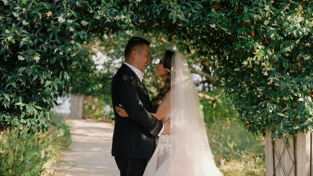 Bride and groom holding each other under a lush jasmine flower arch on a garden path by a Big Sur wedding photographer