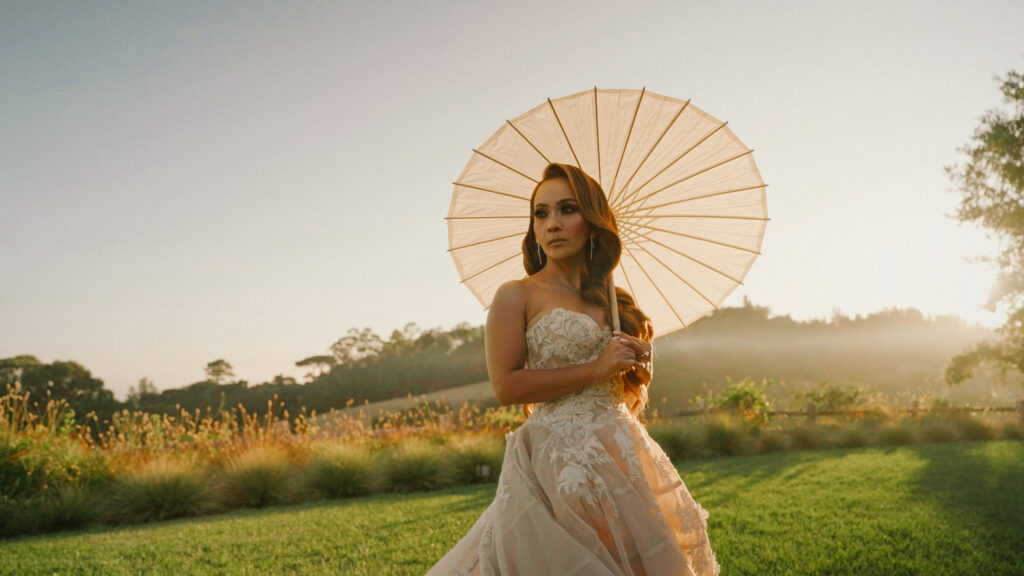 Bride holding a white paper parasol umbrella on a grassy meadow during golden hour captured by a Big Sur wedding photographer