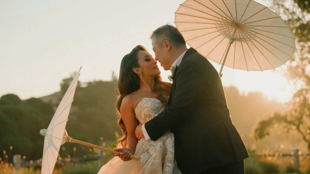 Groom embracing his bride while holding white paper umbrellas on a grassy hill captured by a Big Sur wedding photographer