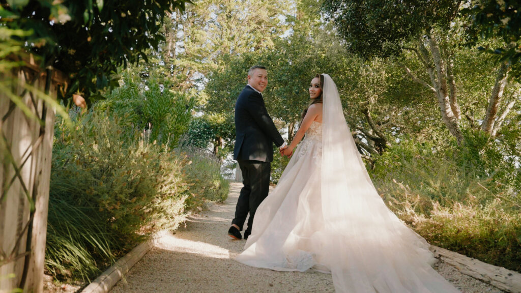 Bride with a long tulle wedding dress train looking back while walking down a garden path with her groom captured by a Big Sur wedding photographer