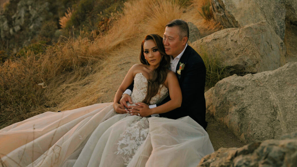 Groom embracing his bride from behind while sitting among rocky cliffs during a sunset Big Sur wedding photoshoot