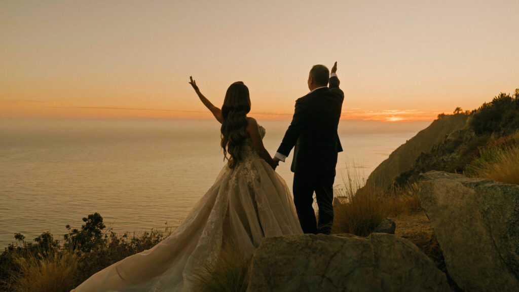 Bride and groom holding hands with raised arms looking at the ocean sunset captured by a Big Sur wedding photographer