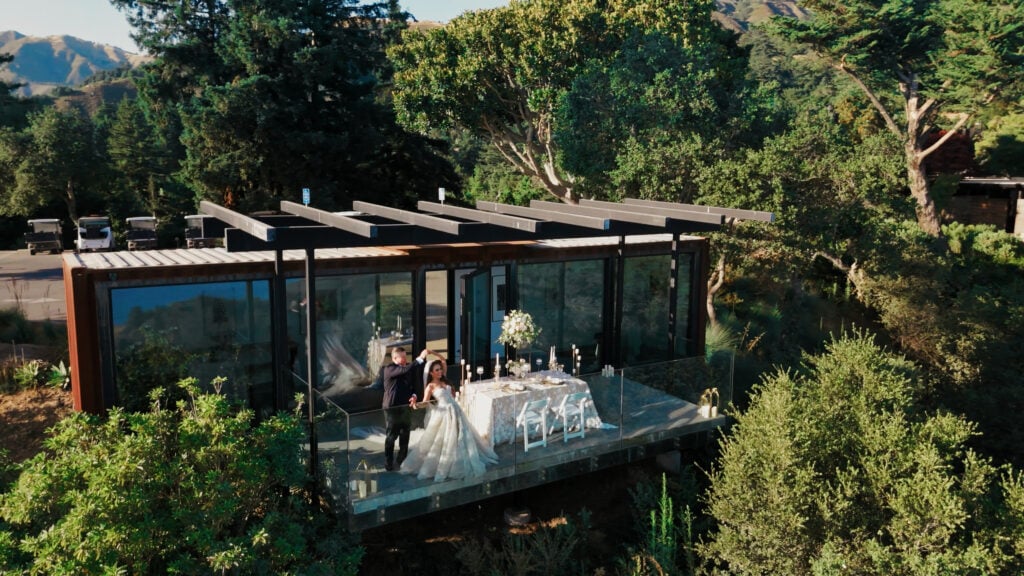 Drone shot of a bride and groom dancing on a glass balcony terrace next to an outdoor reception dinner table by a Big Sur wedding photographer
