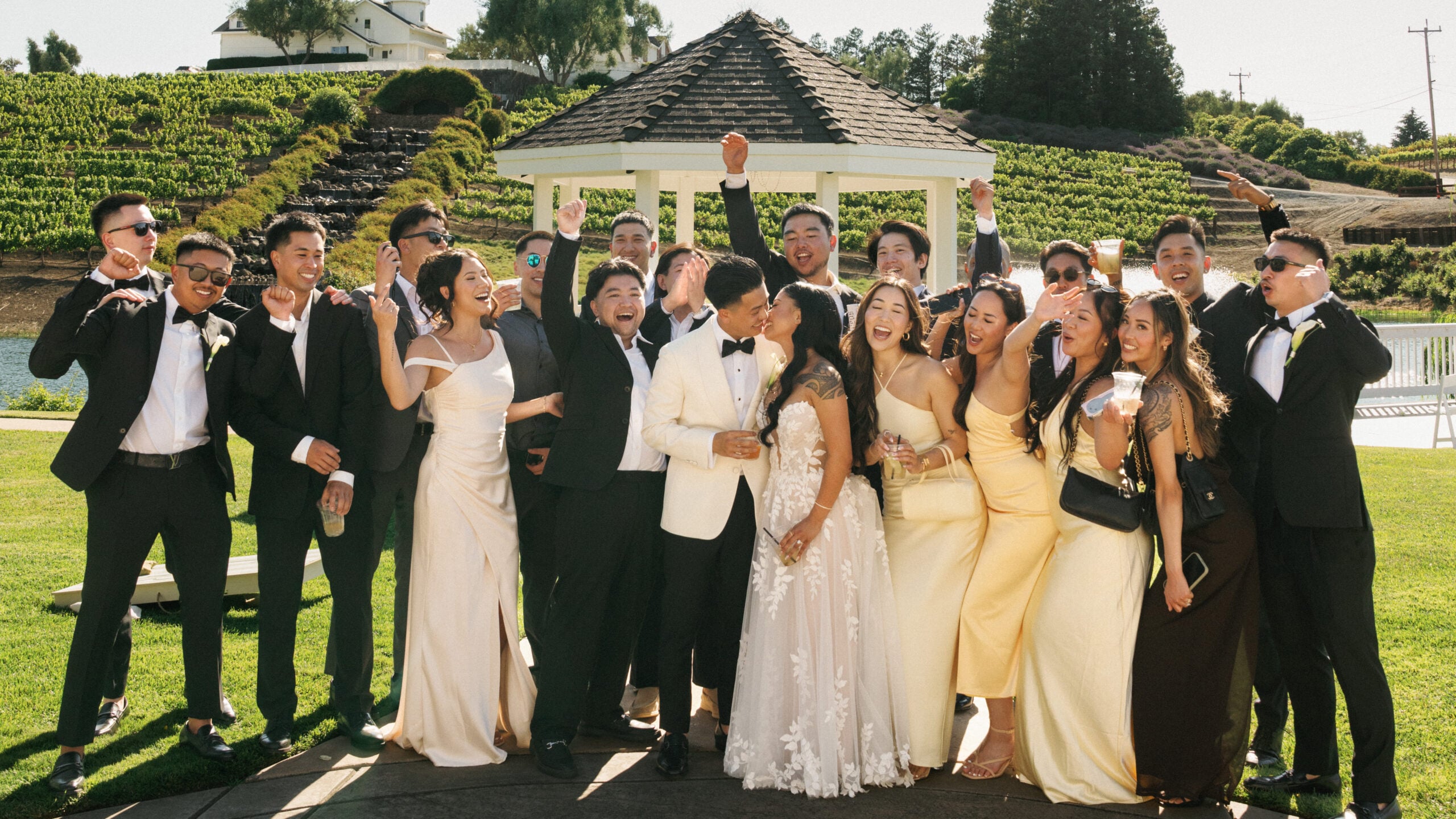 Joyful bride and groom kissing, surrounded by cheering wedding guests and a bridal party in chic champagne yellow dresses and black tuxedos during their lively wedding weekend coverage at a scenic vineyard resort.
