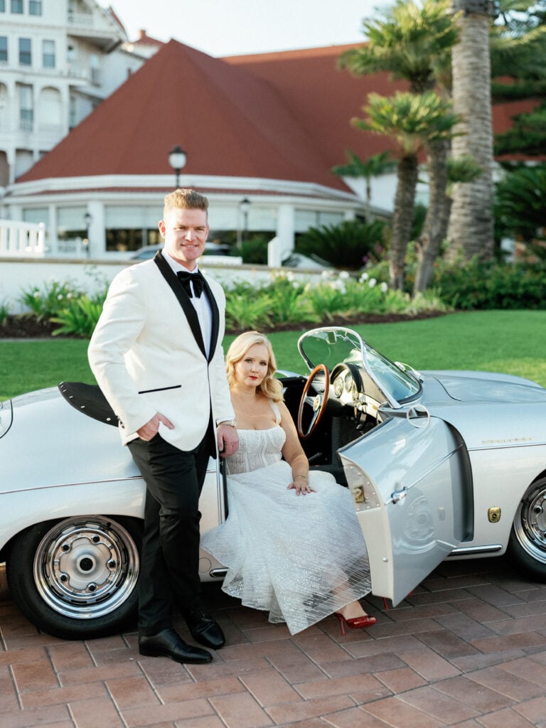 Elegant bride in a sparkly tea-length wedding gown and groom in a white tuxedo jacket posing with a vintage Porsche Speedster, capturing cinematic editorial wedding photography.