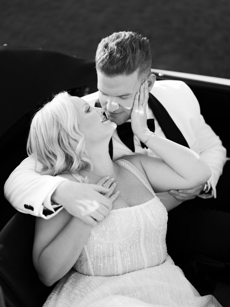 Black and white bride in a beaded wedding gown and groom in a white tuxedo hugging inside a classic car, shot on film by a Palm Springs wedding photographer.