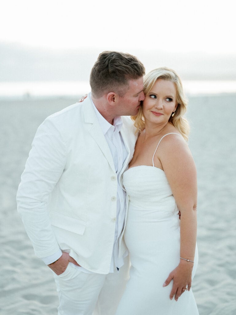 Groom in a white suit kissing his bride's cheek on a sandy beach, shot on film by a Palm Springs wedding photographer.