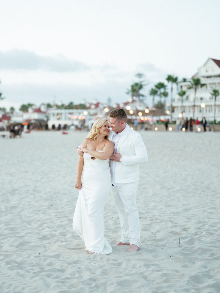 Bride and groom in all-white wedding attire posing on a sandy beach with palm trees and a luxury resort background, captured on film by a Palm Springs wedding photographer.