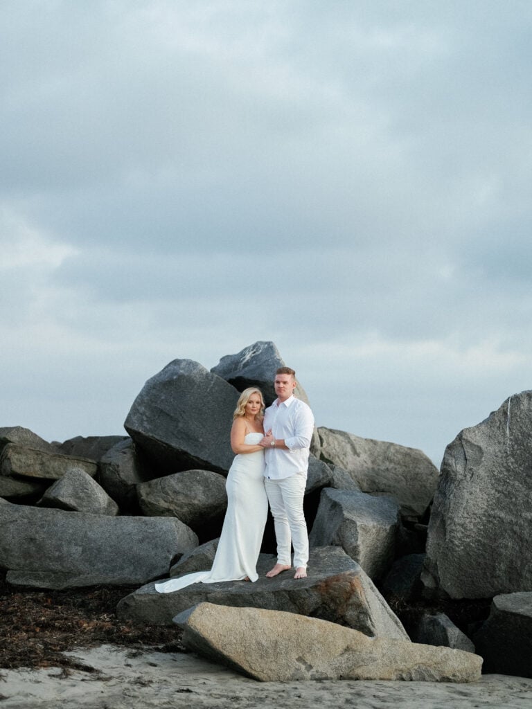 Wide shot of a couple in white attire standing on rocky beach terrain under a cloudy sky, captured on film by a Palm Springs wedding photographer.